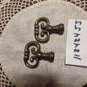 May include: Two antique gold-toned key-shaped drawer pulls with ornate designs. The pulls are on a white crocheted surface with a handwritten note that says "5.75 each".