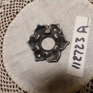 May include: A dark metal, flower-shaped object with a central hole, possibly a decorative element. The object is set on a light-coloured, textured surface, with a tag that reads "112723 A".