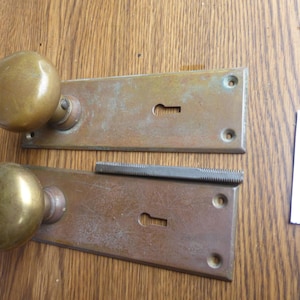 May include: Two brass door knobs attached to rectangular backplates with keyholes, featuring a weathered patina. A metal spindle with a threaded texture is positioned between the two plates. A small white paper tag displays the text 30224 F.