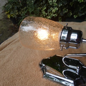 Może przedstawiać: Chromowa lampa ścienna z kloszem z matowego szkła. Lampa ma zakrzywiony wysięgnik i ozdobną podstawę. Światło jest włączone, a żarówka jest widoczna.