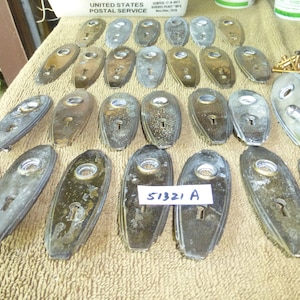 May include: A collection of 25 oval-shaped, antique, metal door plates with keyholes. The plates are in various shades of brass and silver, and some have a tarnished finish. The plates are arranged in rows on a beige, textured surface. A handwritten label with the text "51321 A" is visible in the lower center of the image.