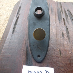 May include: Black oval-shaped door plate with a round brass insert and a single hole at the top. The plate has a D and the number 2221 written on it.