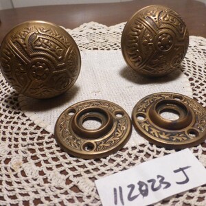 May include: Antique bronze door knobs and matching backplates. The knobs have an ornate, floral design. The backplates are round with a central hole and decorative edges. The image also includes a handwritten tag with the numbers "112823 J".