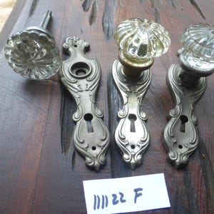 Pode incluir: Três maçanetas de porta antigas com placas de metal ornamentadas e maçanetas de vidro transparente. As maçanetas têm um design decorativo e canelado. As placas de metal têm uma fechadura e volutas intrincadas. O texto "III 22 F" está escrito em um rótulo branco.