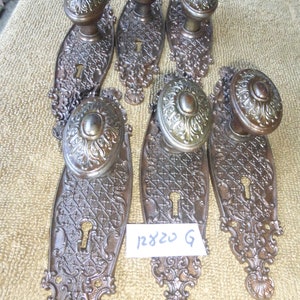 May include: Set of six antique door knobs with ornate designs. Each knob is made of metal and has a keyhole. The knobs are in a dark brown colour with a gold finish. The knobs are arranged in a row on a beige surface.