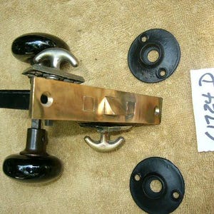 Puede incluir: Una cerradura de puerta de latón con dos pomos de porcelana negra y dos placas de metal negro. La cerradura tiene una cerradura triangular y una manija de palanca. Las placas tienen un agujero para tornillos y un pequeño agujero en el centro. El texto "612219" está escrito en un papel blanco.