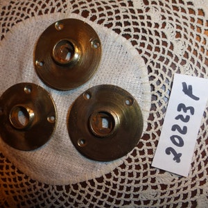 以下が含まれることがあります： 中央に正方形の穴が開いたアンティークな真鍮製のドアノブが3つあります。ノブは、手書きで「202237」と書かれた白いレースのドイリーの上に置かれています。