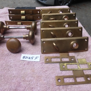 Könnte beinhalten: Eine Sammlung von Türbeschlägen aus Messing im Vintage-Stil, darunter Türknöpfe, Abdeckplatten und Schließbleche. Die Beschläge weisen eine warme, gealterte Patina auf. Ein kleines weißes Etikett mit dem Text "82125 F" ist sichtbar.