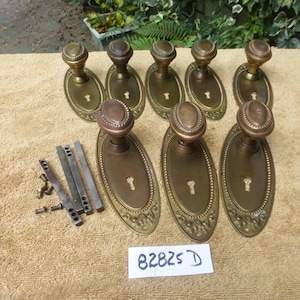 以下が含まれることがあります： アンティーク真鍮製のドアノブとバックプレートのコレクション。ノブは丸みを帯びた上部を持ち、鍵穴と装飾的な縁取りのある楕円形のバックプレートに取り付けられています。「82825 D」のテキストが付いたラベルと取り付け金具が含まれています。