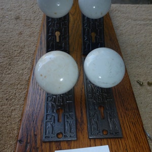 May include: Four antique white porcelain door knobs with ornate black metal plates. The plates have a keyhole and decorative designs. The knobs are mounted on a wooden board with the number "C 02122" written on it.