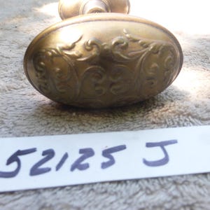 Può includere: Un'antica maniglia per porta in ottone, di forma ovale e con motivi floreali a volute. La maniglia è di un caldo colore dorato e sembra invecchiata. Sotto, un'etichetta bianca riporta i numeri "52125 J" in blu scuro.