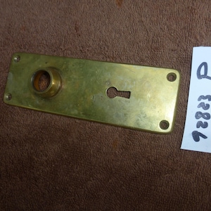 Op de afbeelding: Een rechthoekige messing deurplaat met een sleutelgat en een cirkelvormige opening. De plaat heeft vier schroefgaten en is licht aangetast. Een wit label met de tekst "P 98836" staat naast de plaat.