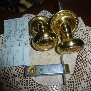 Può includere: Due pomelli per porte dorati con un designo a corda attorno al bordo. I pomelli sono su un pizzo bianco con un chiavistello per porte argentato. Le istruzioni per l'installazione dei pomelli sono visibili sullo sfondo.