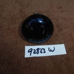 May include: A dark, round glass object with a small loop at the top. The object is black and reflective, with some imperfections on the surface. A white label with the numbers "92823 W" is below the object.