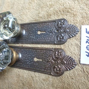 May include: Two door knobs with clear, faceted glass handles and rectangular, dark bronze-toned metal backplates featuring intricate embossed floral and shell patterns, a keyhole, and a small white tag with the handwritten text 41024 F.