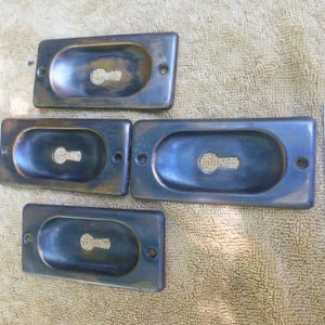 May include: Four antique keyhole escutcheons in a rectangular shape with a rounded oval opening. The escutcheons are made of a dark metal with a worn, aged finish. The escutcheons are arranged in two rows of two, with the top row slightly offset to the left.