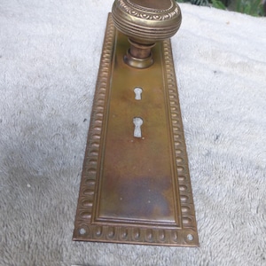 Op de afbeelding: Antieke messing deurklinkset met een ronde knop en rechthoekige achterplaat. De knop heeft een gedetailleerd, gegroefd ontwerp. De achterplaat heeft twee sleutelgaten en een decoratieve rand. Het nummer "52825 J" is gedrukt.