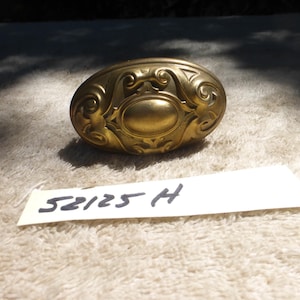 May include: An antique brass oval door knob with ornate scrollwork and a central oval design. The knob is a warm gold color and has a vintage aesthetic. The label below reads "52125 H".