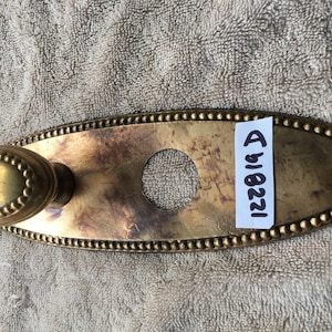 May include: Oval-shaped brass door plate with a decorative knob and a round hole in the center. The plate has a beaded edge and a worn, aged finish. The text "1228/A" is written on a white label attached to the plate.