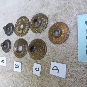 Puede incluir: Una colección de medallones de techo y herrajes antiguos de latón, que incluyen varios tamaños y patrones decorativos. Los artículos están dispuestos sobre una superficie texturizada, con una etiqueta azul que muestra el número "52025 C".
