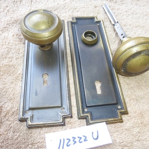 Może przedstawiać: Zestaw antycznych mosiężnych okuć drzwiowych z dziurką od klucza i ozdobną ramką. Okucia obejmują gałkę drzwiową, płytkę drzwiową i klucz. Płytka drzwiowa ma czarne tło z mosiężną ramką.