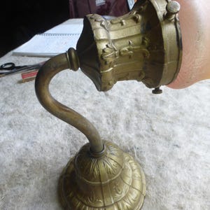 Może przedstawiać: Antyczny kinkiet ścienny z mosiężnym wykończeniem. Kinkiet posiada zakrzywione ramię, dekoracyjną podstawę i szczegółowy uchwyt na klosz. Klosz ma delikatny różowy kolor. Kinkiet jest przeznaczony do montażu na ścianie.