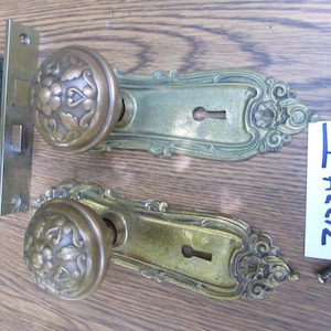 May include: A set of two brass door knobs with ornate floral patterns, attached to matching rectangular backplates with keyholes. The set includes a black metal mortise lock mechanism, a metal spindle, and two screws, with a tag labeled 71224.