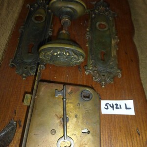 May include: Antique brass door hardware set with a keyhole, key, and a decorative doorknob. The set includes two plates with ornate designs. The label reads "5421 L".