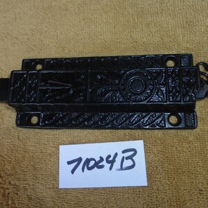 May include: Black cast iron door latch with an ornate design. The latch has a hook on one end and a rectangular plate with a keyhole on the other. The latch is labeled with the number 702413.
