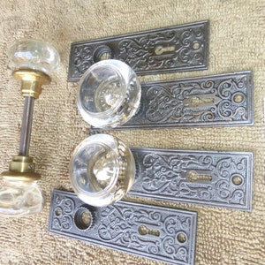 Op de afbeelding: Set van vier antieke deurknopjes met sierlijke metalen platen en heldere glazen knoppen. De platen hebben een decoratief bloemmotief. De knoppen zijn in goede staat en hebben een messing afwerking. De platen zijn gemarkeerd met het nummer "F 22-15".