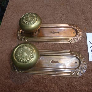 Può includere: Due maniglie antiche in ottone con placche posteriori abbinate. I pomelli sono rotondi con un motivo floreale e le placche sono rettangolari con bordi decorativi. Le serrature sono visibili. Il testo "92823 Y" è su un'etichetta bianca.