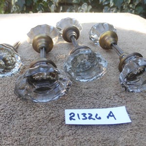 May include: A collection of five clear glass door knobs with brass-colored metal stems and bases. The knobs have a faceted, flower-like design. A small white label with the text "21326 A" is visible.
