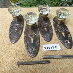May include: Four antique door handle sets with clear glass knobs and ornate, dark metal backplates. Each plate has a keyhole. Two metal rods are also present. The items are arranged on a beige surface.