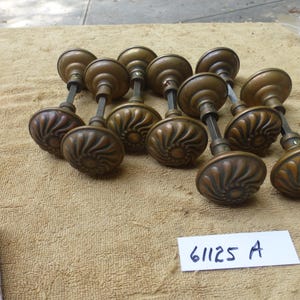 以下が含まれることがあります： 丸いハンドルに詳細なスパイラルデザインが施された、アンティークのブロンズ製ドアノブのコレクション。ノブはベージュ色の表面に配置されています。「61125 A」というテキストの白いラベルが見えます。