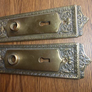 May include: Two rectangular brass door plates featuring ornate embossed scrollwork borders, a central circular knob opening, and a keyhole cutout. A small white paper tag to the right displays the handwritten text 30324 L in blue ink.