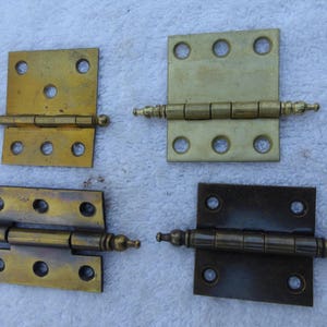 May include: Four antique brass hinges with decorative finials. The hinges are all the same size and shape, but have different finishes. Two are a bright brass, one is a darker brass, and one is a bronze color.