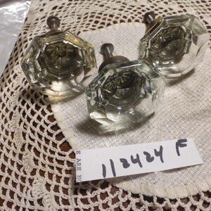 May include: Three clear glass door knobs with silver metal accents. The knobs are on a white crocheted doily. A white tag with the number "113424 F" is visible.