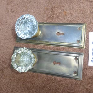 Op de afbeelding: Twee antieke deurklinksets. Elke set bevat een heldere, facetgeslepen glazen knop en een rechthoekige metalen achterplaat met een sleutelgat. De achterplaten hebben een messingkleurig frame en een zilverkleurig midden. Een wit label met het nummer "92823 5" is zichtbaar.