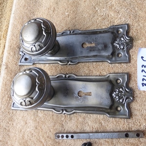 May include: Two antique silver door knobs with keyholes and decorative plates. The plates have a floral design. The knobs are round with a raised center. The plates are attached to a metal bar with four holes. The image also includes a handwritten tag with the number 32123 C.