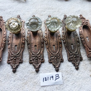 May include: Set of six antique door knobs with ornate brass plates and clear glass knobs. The plates have a keyhole and intricate floral designs. The knobs are faceted and have a slight greenish tint. The set is labeled "12119 B".