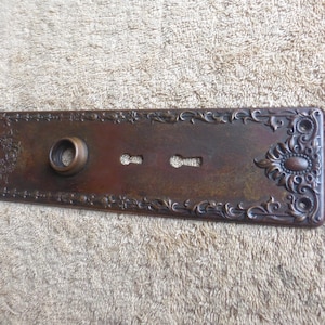 May include: A rectangular, antique bronze door plate with ornate floral and scrollwork detailing. It features a circular knob opening, two keyhole slots, and a decorative oval design. The text "N 42.92.1" is visible on a white label.