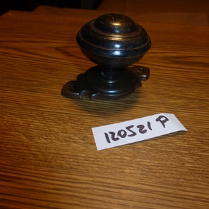 May include: Black metal door knob with a round, fluted handle and a decorative base. The knob is on a wooden surface with a white tag that says "P 120521".