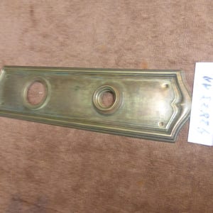 Könnte beinhalten: Rechteckige Türplatte aus Messing mit zwei kreisförmigen Löchern und dekorativen Kanten. Die Platte hat ein angelaufenes Aussehen, was auf Alter hindeutet. Ein weißes Etikett mit dem Text "42823 1A" ist sichtbar.