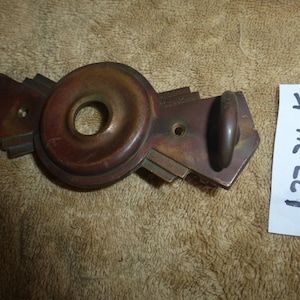 May include: An antique bronze-colored door handle with a circular center and geometric side details. The handle has a small, rounded knob on one end. A white label with the text "1.27.24 K" is visible.