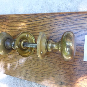 May include: Brass door knob set with a round knob and a backplate. The knob is a traditional style with a smooth finish. The backplate has three screws. The set is in good condition and has a vintage look.