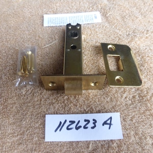 Peut inclure: Un ensemble de loquet de porte en laiton avec une plaque de frappe, des vis et des instructions. L'ensemble est étiqueté avec le numéro "112623 A".