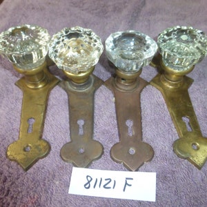 May include: Four antique brass door knobs with clear glass crystal knobs. The knobs have a keyhole and a decorative design at the bottom. The knobs are arranged in a row on a purple surface. 81121 F