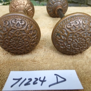 May include: Two round bronze door knobs featuring an intricate embossed floral and geometric pattern across the surface. Each knob has a central circular design surrounded by scrolling motifs. A white paper tag with the handwritten text 71224 D.