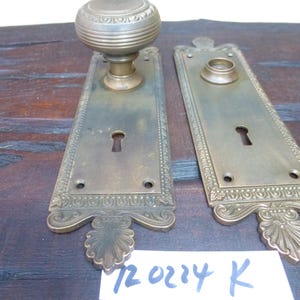 May include: Two antique brass door plates with ornate designs and keyholes. The plates have a decorative border and a floral design at the bottom. The plates are on a wooden surface with the text "720214 K" written on a white piece of paper.