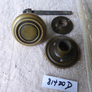 May include: Brass door knob set with a round, decorative knob and two round plates. The knob has a concentric circle design. The plates have a raised lip around the edge. The set is in good condition with some signs of wear.